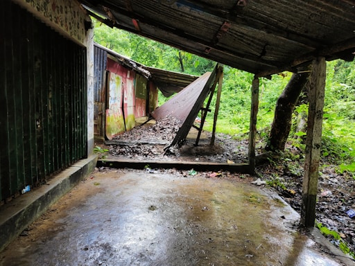 A dilapidated and abandoned shelter with a slanted tin roof, surrounded by dense greenery. The area is wet and littered with fallen leaves and twigs. The walls of the structure are discolored, featuring faded graffiti and moss growth.