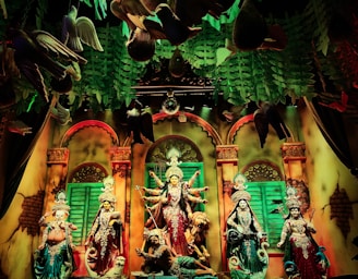 A vibrant and colorful scene of Hindu deities in a richly decorated setting. The figures are adorned with intricate clothing and jewelry. The background features arches and lush green foliage, while birds appear to be suspended in the air. The atmosphere suggests a festive or religious celebration.