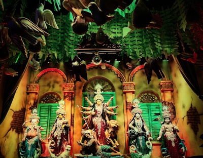 A vibrant and colorful scene of Hindu deities in a richly decorated setting. The figures are adorned with intricate clothing and jewelry. The background features arches and lush green foliage, while birds appear to be suspended in the air. The atmosphere suggests a festive or religious celebration.