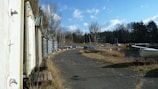 A row of weathered white sheds with metal roofs line a cracked asphalt path. To the right, several small boats are positioned along a grassy shoreline next to a calm body of water. Tall, leafless trees and a dense evergreen forest form a backdrop under a sky with fluffy white clouds.
