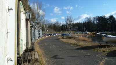 A row of weathered white sheds with metal roofs line a cracked asphalt path. To the right, several small boats are positioned along a grassy shoreline next to a calm body of water. Tall, leafless trees and a dense evergreen forest form a backdrop under a sky with fluffy white clouds.