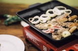 On a tabletop grill, a variety of foods are being cooked, including circular onion slices, sausages, thin strips of meat, and bacon. The grill is placed on a red portable stove with a wooden table and a white plate nearby.