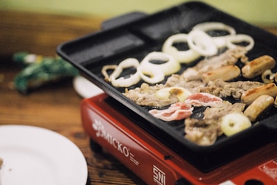 On a tabletop grill, a variety of foods are being cooked, including circular onion slices, sausages, thin strips of meat, and bacon. The grill is placed on a red portable stove with a wooden table and a white plate nearby.