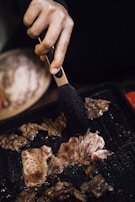 A hand holding black tongs is flipping slices of cooking meat on a grill pan. The meat appears to be sizzling, with visible juices and a slightly browned exterior.
