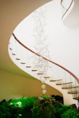 A spiral staircase with a wooden railing curves gracefully above a lush green indoor garden. The focal point is a delicate art installation featuring interconnected rods and translucent spheres suspended in the center of the staircase.