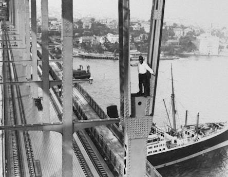 A man is standing on a steel beam high above the ground, with a view of an urban waterfront area in the background. The scene includes a large bridge structure with metal rivets, railway tracks, and a train visible. Below, a large ship is docked near the shore, alongside various buildings and greenery.
