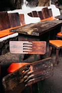 A wooden table and chairs set with distinctive rustic, dark wood grain is placed indoors. The chairs feature orange cushions, and the table is surrounded by a stone wall with white outlines.