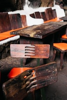 A wooden table and chairs set with distinctive rustic, dark wood grain is placed indoors. The chairs feature orange cushions, and the table is surrounded by a stone wall with white outlines.