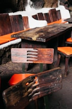 A wooden table and chairs set with distinctive rustic, dark wood grain is placed indoors. The chairs feature orange cushions, and the table is surrounded by a stone wall with white outlines.
