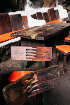 A wooden table and chairs set with distinctive rustic, dark wood grain is placed indoors. The chairs feature orange cushions, and the table is surrounded by a stone wall with white outlines.