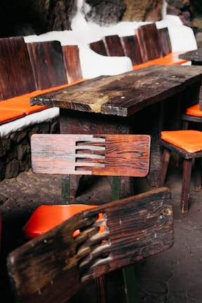 A wooden table and chairs set with distinctive rustic, dark wood grain is placed indoors. The chairs feature orange cushions, and the table is surrounded by a stone wall with white outlines.