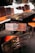 A wooden table and chairs set with distinctive rustic, dark wood grain is placed indoors. The chairs feature orange cushions, and the table is surrounded by a stone wall with white outlines.