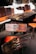 A wooden table and chairs set with distinctive rustic, dark wood grain is placed indoors. The chairs feature orange cushions, and the table is surrounded by a stone wall with white outlines.