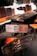A wooden table and chairs set with distinctive rustic, dark wood grain is placed indoors. The chairs feature orange cushions, and the table is surrounded by a stone wall with white outlines.