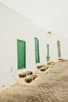 A row of white buildings with vibrant green doors set against a cobblestone street creates a rustic and quaint atmosphere. The buildings show signs of wear, with peeling paint and a textured surface. Small stone steps lead up to each door, adding to the charming and aged aesthetic.
