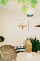 A cozy indoor space with a white rounded seating area featuring blue and white striped cushions. A potted plant with lush green leaves adds a natural touch. A unique art piece depicting fish with geometric patterns is framed on the wall. A decorative object resembling a pinecone is prominently displayed on a white pedestal table. Soft natural lighting creates a serene and balanced atmosphere.