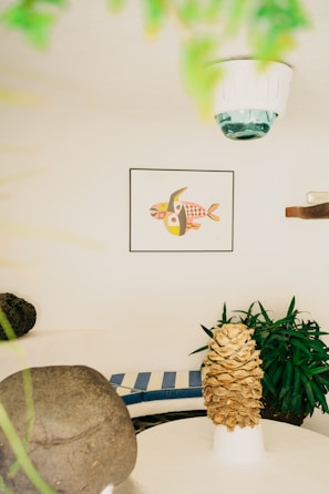 A cozy indoor space with a white rounded seating area featuring blue and white striped cushions. A potted plant with lush green leaves adds a natural touch. A unique art piece depicting fish with geometric patterns is framed on the wall. A decorative object resembling a pinecone is prominently displayed on a white pedestal table. Soft natural lighting creates a serene and balanced atmosphere.