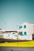 A bright, white building with vibrant turquoise shutters and accents stands under a clear, blue sky. Surrounding it are tropical plants and a low stone wall. In the foreground, a yellow object, possibly a boat or barrier, partially obstructs the bottom of the view.