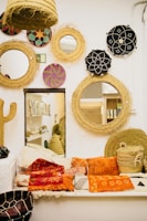An interior space featuring a collection of round woven wall decorations and mirrors, complemented by an assortment of colorful textiles in orange and red hues. The wall displays a variety of woven baskets with intricate patterns, including some vibrant ethnic designs. Below, a seating area is adorned with numerous cushions and throws, creating a cozy and inviting atmosphere.