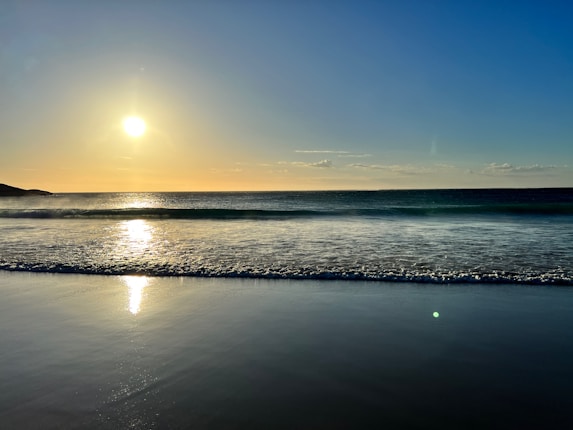 A tranquil beach scene at sunset with the sun low on the horizon reflecting off gentle waves. The sky is mostly clear with a few scattered clouds, casting a warm glow over the ocean and wet sand.