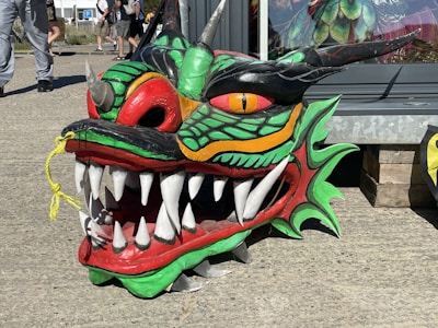 A colorful dragon head made of vibrant materials is positioned on a concrete surface. It features large, sharp white teeth, vivid green and red scales, and a prominent yellow eye. The background includes a glass storefront and a wooden pallet.
