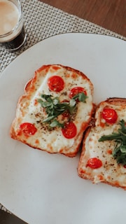 Two slices of toasted bread topped with melted cheese, cherry tomatoes, and green herbs sit on a white plate. A small glass of coffee is placed to the left of the plate on a woven placemat.