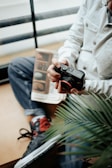 A person sits casually holding a camera, with a magazine or book on their lap. They are wearing casual clothing, including jeans and stylish sneakers with red accents. A green plant is partially visible in the foreground, adding a touch of nature to the scene.