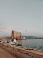 A coastal industrial scene with a large set of silo structures dominating the background. Two small boats are moored in the foreground, while a larger cargo ship is docked near the silos. The sky is overcast, casting a muted, serene atmosphere over the water.