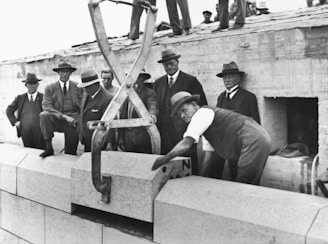 A group of men in early 20th-century formal attire, including suits and hats, are overseeing a construction operation. One man is actively placing a large stone block using a mechanical lifting device while others observe.
