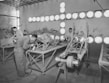 Several individuals are working in a workshop on assembling or painting metal frames. The room is illuminated by multiple circular lights, creating a bright atmosphere. The workers are dressed in overalls, suggesting an industrial or manufacturing setting.