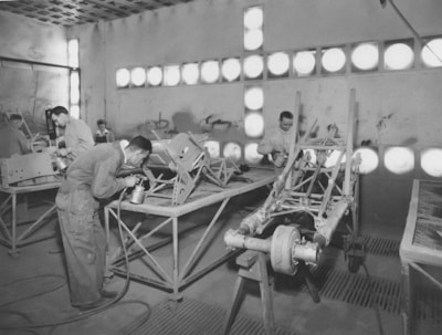 Several individuals are working in a workshop on assembling or painting metal frames. The room is illuminated by multiple circular lights, creating a bright atmosphere. The workers are dressed in overalls, suggesting an industrial or manufacturing setting.