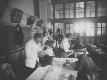 A black and white photograph depicts a room filled with several people, including children and adults, engaging in what appears to be a medical examination or vaccination. The setting resembles a classroom or clinic, with a long table where children are being attended to by adults. The lighting is bright in the background, and there's a sense of orderliness and attention to the process at hand.