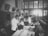 A black and white photograph depicts a room filled with several people, including children and adults, engaging in what appears to be a medical examination or vaccination. The setting resembles a classroom or clinic, with a long table where children are being attended to by adults. The lighting is bright in the background, and there's a sense of orderliness and attention to the process at hand.