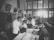 A black and white photograph depicts a room filled with several people, including children and adults, engaging in what appears to be a medical examination or vaccination. The setting resembles a classroom or clinic, with a long table where children are being attended to by adults. The lighting is bright in the background, and there's a sense of orderliness and attention to the process at hand.