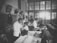 A black and white photograph depicts a room filled with several people, including children and adults, engaging in what appears to be a medical examination or vaccination. The setting resembles a classroom or clinic, with a long table where children are being attended to by adults. The lighting is bright in the background, and there's a sense of orderliness and attention to the process at hand.