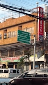 An urban street scene with a building in the background featuring a bank sign. Several green directional road signs indicate directions to Talamban, Cebu City, and Consolacion. Various business signs are visible, including one for Color Options Marketing. There is a parked truck and a car in the foreground. A bundle of overhead wires hangs above the street.