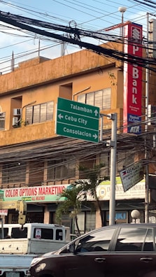 An urban street scene with a building in the background featuring a bank sign. Several green directional road signs indicate directions to Talamban, Cebu City, and Consolacion. Various business signs are visible, including one for Color Options Marketing. There is a parked truck and a car in the foreground. A bundle of overhead wires hangs above the street.
