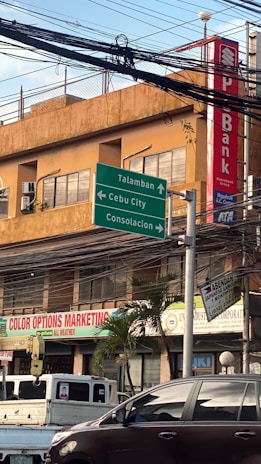 An urban street scene with a building in the background featuring a bank sign. Several green directional road signs indicate directions to Talamban, Cebu City, and Consolacion. Various business signs are visible, including one for Color Options Marketing. There is a parked truck and a car in the foreground. A bundle of overhead wires hangs above the street.