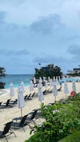 A sandy beach with multiple rows of closed white umbrellas and black sun loungers. The ocean is in the background, with waves gently crashing on the shore. Lush greenery is in the foreground, and scattered small vacation cottages are visible along the beach. The sky is overcast with clouds.