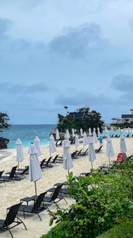 A sandy beach with multiple rows of closed white umbrellas and black sun loungers. The ocean is in the background, with waves gently crashing on the shore. Lush greenery is in the foreground, and scattered small vacation cottages are visible along the beach. The sky is overcast with clouds.
