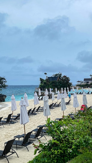 A sandy beach with multiple rows of closed white umbrellas and black sun loungers. The ocean is in the background, with waves gently crashing on the shore. Lush greenery is in the foreground, and scattered small vacation cottages are visible along the beach. The sky is overcast with clouds.