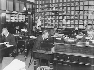 A black and white photograph of an early 20th-century office setting featuring several men working at wooden desks. The room is filled with shelves of files and documents, typewriters, and organization boxes. The men are dressed in formal attire, indicative of the period, and are engrossed in tasks such as writing and typing.