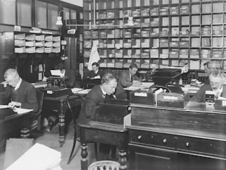 A black and white photograph of an early 20th-century office setting featuring several men working at wooden desks. The room is filled with shelves of files and documents, typewriters, and organization boxes. The men are dressed in formal attire, indicative of the period, and are engrossed in tasks such as writing and typing.