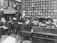 A black and white photograph of an early 20th-century office setting featuring several men working at wooden desks. The room is filled with shelves of files and documents, typewriters, and organization boxes. The men are dressed in formal attire, indicative of the period, and are engrossed in tasks such as writing and typing.