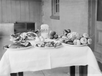 A black and white photograph of a table set with various foods including large cuts of meat, a pitcher of milk, eggs, and a variety of fruits and vegetables such as cauliflower, apples, and turnips. These items are displayed on plates and arranged neatly across a tablecloth.