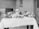 A black and white photograph of a table set with various foods including large cuts of meat, a pitcher of milk, eggs, and a variety of fruits and vegetables such as cauliflower, apples, and turnips. These items are displayed on plates and arranged neatly across a tablecloth.