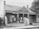 A historical black and white photo of a brick building with a sign reading 'Baby Clinic'. Several women in vintage nursing uniforms, along with infants, are standing and seated outside on the steps. The architecture features white wooden trim and a decorative fence encloses the front.