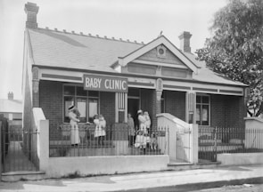 A historical black and white photo of a brick building with a sign reading 'Baby Clinic'. Several women in vintage nursing uniforms, along with infants, are standing and seated outside on the steps. The architecture features white wooden trim and a decorative fence encloses the front.
