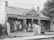 A historical black and white photo of a brick building with a sign reading 'Baby Clinic'. Several women in vintage nursing uniforms, along with infants, are standing and seated outside on the steps. The architecture features white wooden trim and a decorative fence encloses the front.