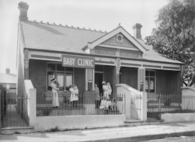 A historical black and white photo of a brick building with a sign reading 'Baby Clinic'. Several women in vintage nursing uniforms, along with infants, are standing and seated outside on the steps. The architecture features white wooden trim and a decorative fence encloses the front.